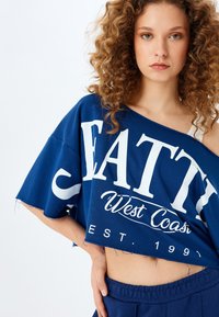 Marinblå kortärmad T-shirt med vit text som läser "SEATTY West Coast EST. 1991", med avslappnad passform och slitna kanter. Mjuk tygkänsla.