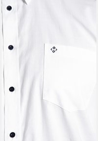 Weißes Baumwollhemd mit Knopfleiste, ausgestattet mit einer aufgesetzten Tasche und einem gestickten Design. Dunkle Knöpfe bilden einen Kontrast zu dem hellen Stoff.