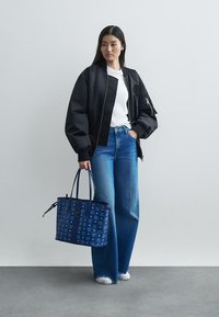 Veľký čierny bomber kabát, biele tričko, široké modré džínsy a vzorovaná námornícka taška. Model stojí na sivej podlahe.