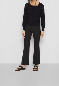 Svart stickad tröja med fyrkantig halsringning och ögondetaljer på ärmarna, i kombination med svarta utsvängda byxor och svarta sandaler med rosettdekorationer.