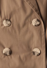 Cappotto doppiopetto in tessuto marrone chiaro, con grandi bottoni beige, texture liscia e leggero lucido.