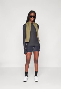 Sportinis aprangos derinys: alyvuogių žalios spalvos liemenė dėvima ant juodos ilgomis rankovėmis palaidinės, juodi šortai ir bepirštinės pirštinės, derinami su akiniais nuo saulės ir batais.