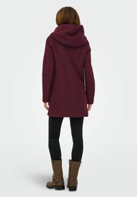 Manteau bordeaux avec une grande capuche, en tissu texturé, à manches longues et coutures verticales. Associé à un pantalon noir skinny et des bottines brunes.