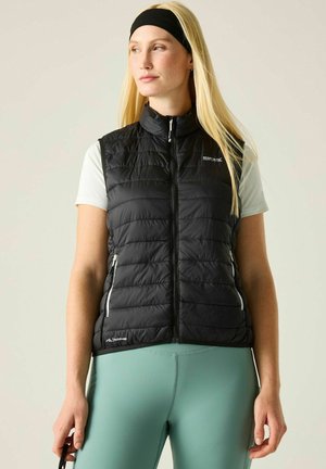 Vrouw met lang blond haar, die een zwart doorgestikt vest, wit shirt met korte mouwen, turquoise leggings en een zwart hoofdbandje draagt, staand en naar rechts kijkend.