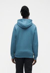 Zils kokvilnas džemperis ar kapuci, kurai ir aukla, ribas aproces un jostasvietas malas. Gluda tekstūra un minimāls dizains, redzams no aizmugures.