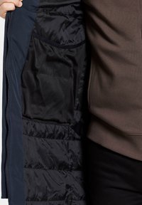 Marinblå ytterkläder med svart, vadderad inredning och nätfickor. Har foder med kviltad struktur och dragkedjestängning längs fronten och fickkanterna.