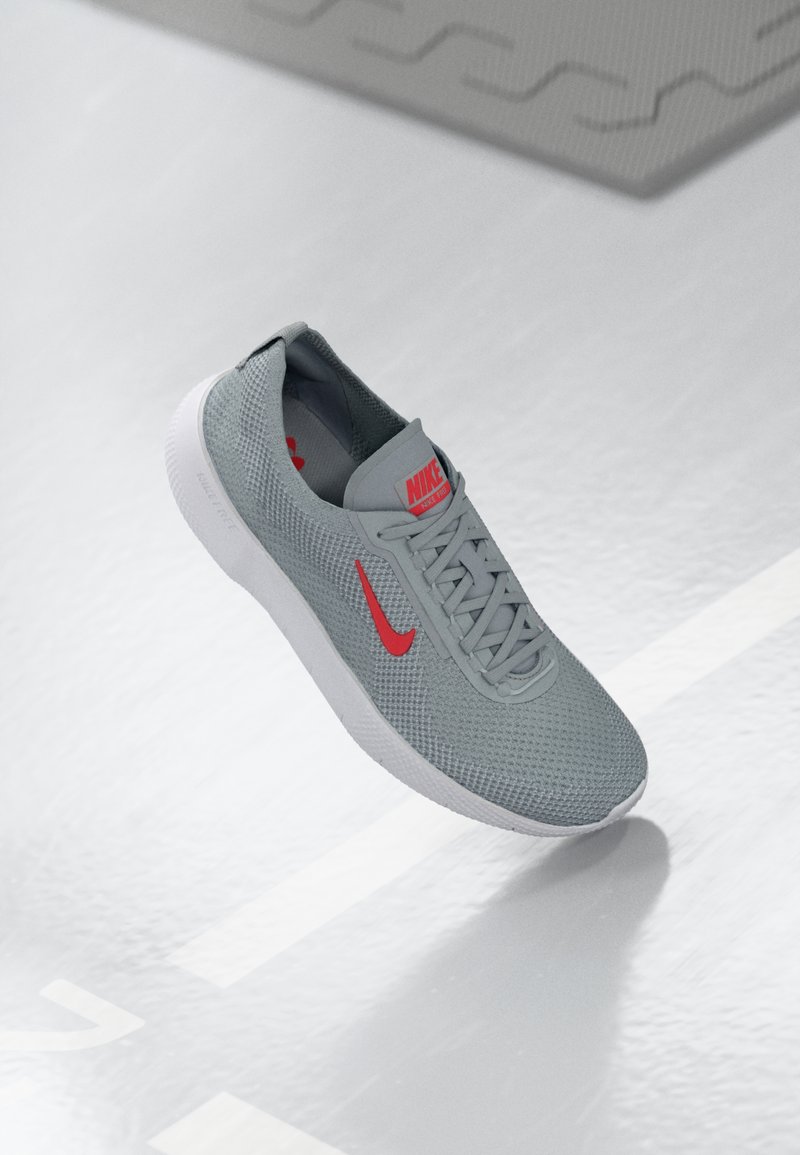 Siva atletska tenisica s teksturiranim mrežastim gornjim dijelom, crvenim Nike logom i bijelim potplatom. Ima zaobljeni vrh i dizajn s vezanjem.