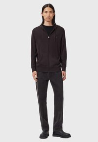 Man som bär en svart zip-up hoodie, svart t-shirt, mörkgrå jeans och svarta stövlar, stående mot en enkel ljus bakgrund.