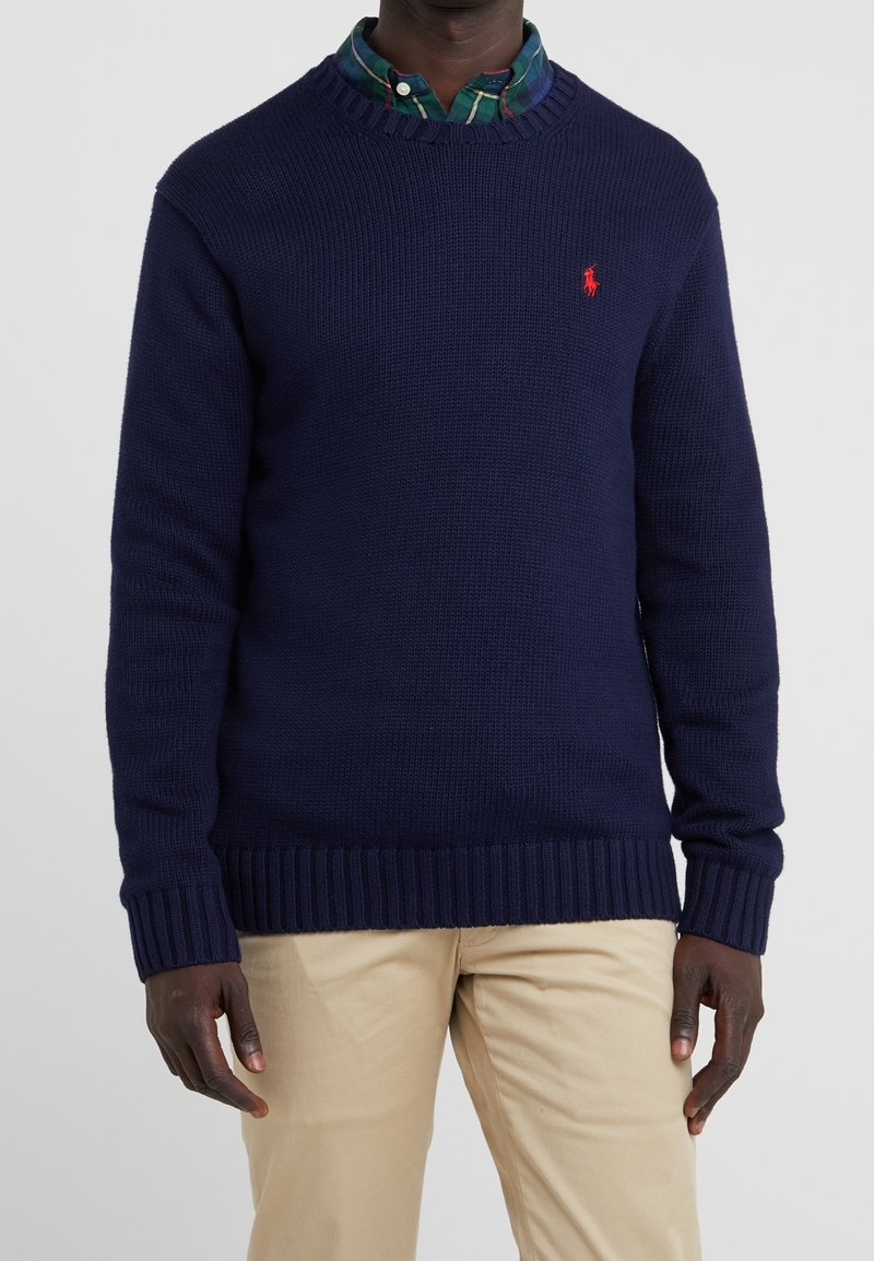 Homme portant un pull en maille bleu marine sur une chemise à carreaux verte et un pantalon beige, debout devant un fond gris uni.