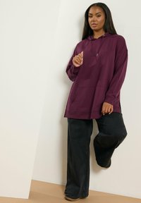Sudadera burdeos de gran tamaño con cordón ajustable, bolsillo frontal y aberturas laterales; combinada con pantalones anchos negros y zapatos negros.