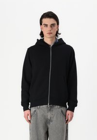 Schwarze Zip-Hoodie aus weichem Stoff, mit einer doppellagigen Kapuze, frontseitigen Taschen und gerippten Bündchen. Getragen über gestreiften grauen Hosen.