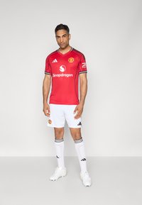 Camisa vermelha do Manchester United com detalhes em preto, apresentando um decote em V, logotipo e texto de patrocínio. Combinada com calções brancos e meias listradas.