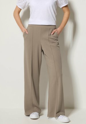 Donna che indossa pantaloni beige a gamba larga con cuciture frontali, mani nelle tasche, sneakers bianche e una maglietta bianca a maniche corte con dettagli traforati.