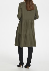 Olive zieleń sukienka z długimi rękawami, warstwowy fason, miękka tkanina; w połączeniu z dopasowanymi czarnymi spodniami i czarnymi szpilkami.