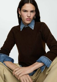 Pulover tricotat maro cu guler și manșete din denim, având accente albastre. Modelul are părul lung și este așezat cu mâinile împreunate.
