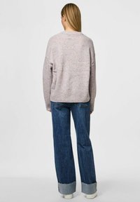 Mujer con cabello largo y rubio lleva un suéter de color claro con manchas, jeans azules de pernera ancha y zapatos blancos, de pie de espaldas contra un fondo liso.