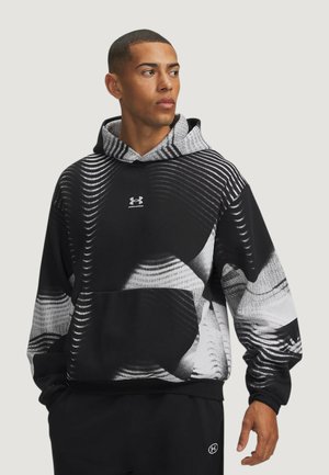 Uomo che indossa una felpa con cappuccio Under Armour a motivo bianco e nero, guarda di lato con le mani lungo i fianchi.