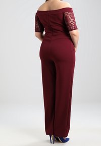 Bordeauxroter Jumpsuit mit schulterfreiem Schnitt, Spitzensäumen an den Ärmeln, eng anliegendem Oberteil, geraden Beinen und Reißverschluss auf der Rückseite. Dazu blaue High Heels.