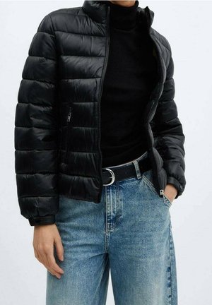 Persoon in een zwarte gewatteerde jas over een zwart coltrui en lichtblauwe denimjeans met een zwarte riem, hand in de zak.