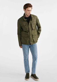 Zielona oliwkowa kurtka z wieloma kieszeniami z przodu, wysokim kołnierzem i lekkim materiałem. W połączeniu z jasnoniebieskimi dżinsami i ciemnymi sneakersami.