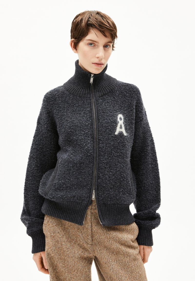 Pull à zip bleu marine avec col haut, tissu texturé, bords côtes aux poignets et une lettre 'A' blanche brodée sur le côté gauche.