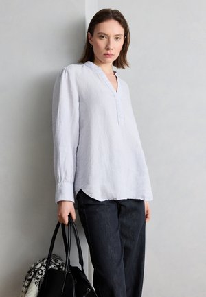 Fehér hosszú ujjú blúzt és sötét farmert viselő nő áll a falnak támaszkodva, kezében fekete kézitáska mintás belsővel.