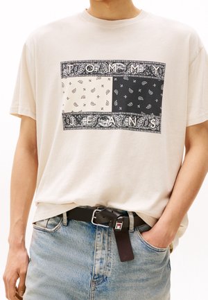T-shirt en coton crème avec un imprimé bandana noir et beige, bordé de motifs décoratifs, avec le texte « TOMMY JEANS » au centre.