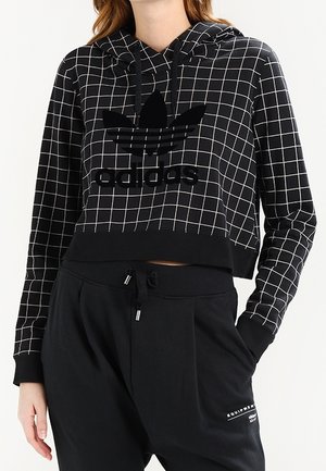 Femme portant un sweat à capuche Adidas noir avec motif à carreaux blanc et un pantalon de jogging noir avec cordon et détails de poches.