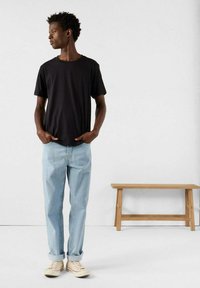 T-shirt nera in cotone con maniche corte, jeans in denim azzurro chiaro e sneakers beige. Il modello è in piedi con le mani in tasca su uno sfondo neutro.