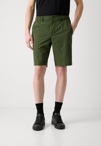 Shorts verts en matériau léger, dotés d'une taille élastique, de poches avant et d'une coupe ajustée. Portés avec des baskets noires et des chaussettes.