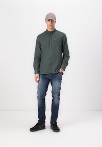 Cămașă cu butoni de culoare verde închis, cu un buzunar pe piept, purtat�ă cu blugi albaștri și adidași negri. O casca bej completează ținuta.