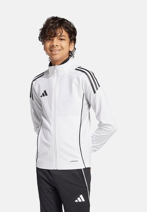 Smilende dreng med krøllet hår iført en hvid Adidas lynjakke med sorte striber og sorte Adidas bukser, står mod en ensfarvet baggrund.