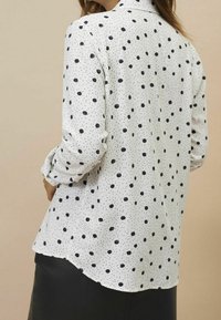 Weiße Bluse mit marineblauen Punkten, langen Ärmeln und abgerundetem Saum. Glattes Material mit feiner gepunkteter Textur verleiht dem Design-detail.