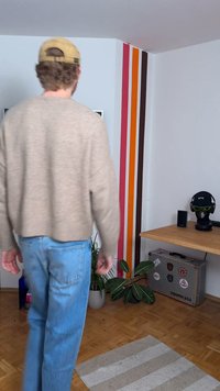 Bež pleteni pulover, svetlo modri džins, oblečena rastlina, lesena miza, črne slušalke na stojalu, pisane stene z obarvanimi črtami, teksturirana preproga.