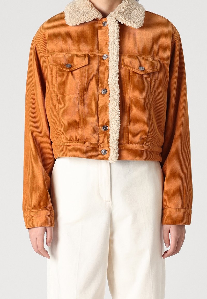 Giacca corta in velluto a coste arancione con morbido colletto in finta pelliccia beige, due tasche sul petto e bottoni automatici argento. Dettaglio del tessuto strutturato visibile.