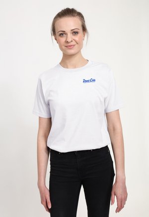 Kvinde med lysebrunt hår sat tilbage iført en hvid Pepsi-Cola t-shirt og sorte bukser, står mod en enkel lys baggrund.