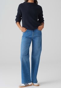 Personne portant un pull en maille noir, un jean large bleu et des baskets blanches à rayures noires, debout les mains dans les poches.