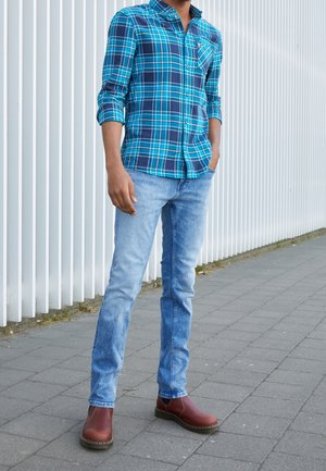 Mann trägt blau kariertes Hemd, hellblaue Jeans und braune Stiefel, steht auf grau gepflastertem Boden neben einem weißen vertikalen Zaun.