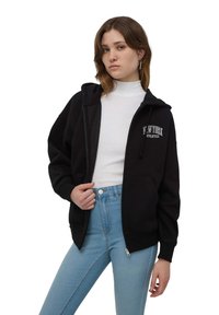 Felpa nera con zip e cappuccio regolabile, con la scritta "New York Athletics" ricamata in bianco. Indossata sopra un dolcevita bianco e dei jeans azzurri chiari.