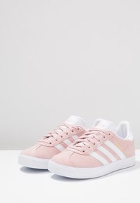 Rosa mockasiner med vita gummisulor, med tre vita ränder och en gul "Gazelle"-logotyp på sidan.