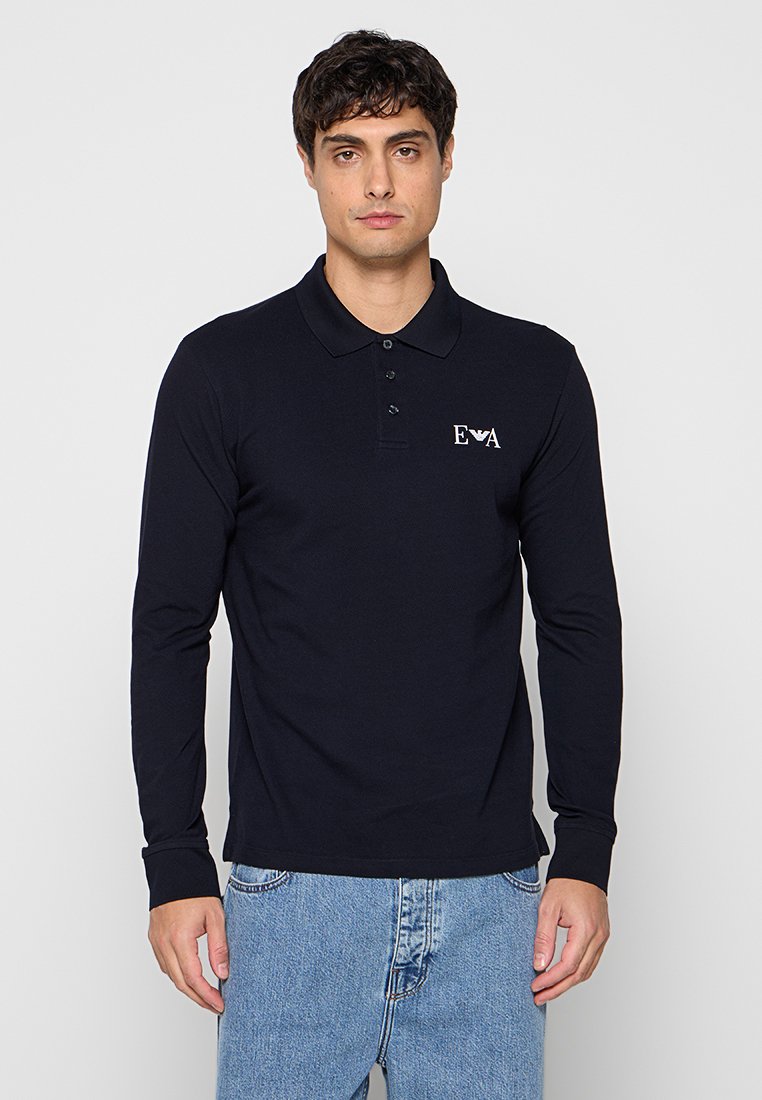 Hombre con una camiseta polo negra de manga larga con el logo "EVA" en el pecho y vaqueros azul claro, de pie frente a un fondo liso.
