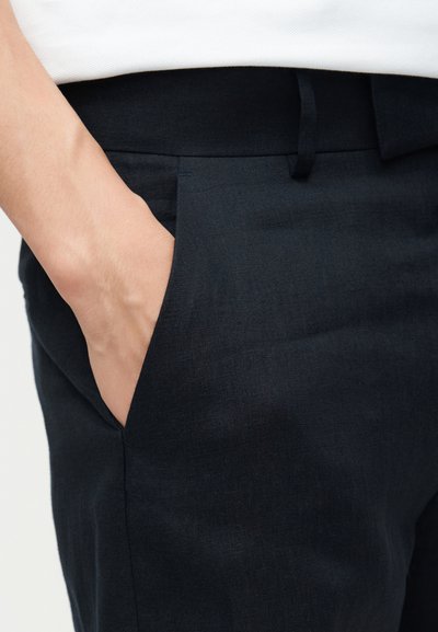 Χέρι με ανοιχτόχρωμο δέρμα που έχει τοποθετηθεί στην μπροστινή τσέπη σκούρων ναυτικών παντελονιών, που φοριούνται με λευκό πουκάμισο.