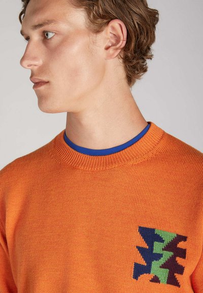 Oranger gestrickter Pullover mit Rundhalsausschnitt und blauer Unterhemden. Besitzt ein mehrfarbiges geometrisches Muster im linken Brustbereich.