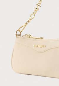 Μπεζ δερμάτινο τσαντάκι χειρός με καμπυλωτή κορυφή, χρυσές μεταλλικές λεπτομέρειες και λουρί αλυσίδας με εναλλασσόμενους κρίκους και διακοσμητικό στοιχείο.