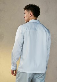 Camisa de manga larga azul claro con una textura suave, puños abotonados y un collar de punta. Presenta un detalle de yugo en la parte posterior para un diseño estructurado.