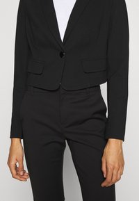 Personne portant un blazer noir ajusté à un seul bouton et un pantalon noir assorti sur un haut blanc, debout avec les bras détendus.