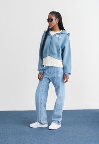 Moletom azul cortado com bolsos frontais, usado sobre uma camisa branca; jeans listrados verticais em azul claro; tênis brancos; óculos de sol azuis.