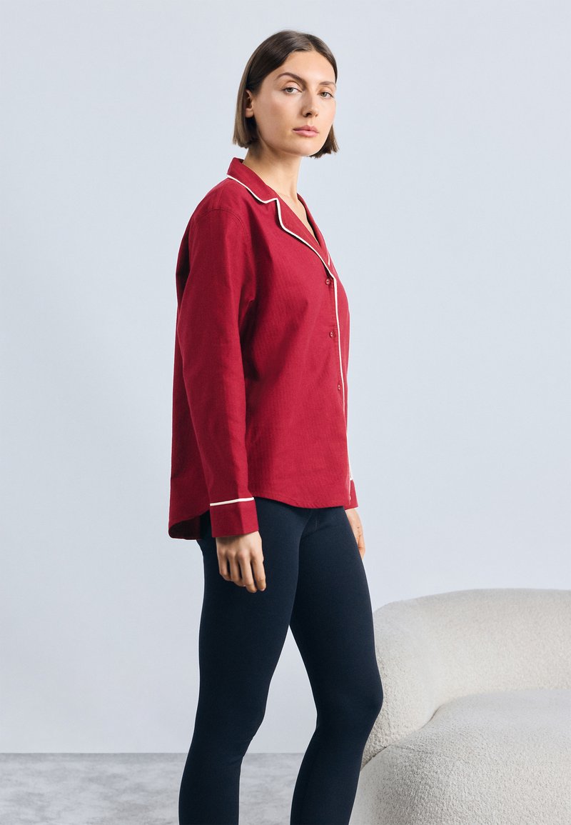 Sarkana krekls ar baltiem apmales akcentiem, saskaņots ar tumši ziliem legingiem. Krekls ir brīvs un ar nedaudz strukturētu audumu.