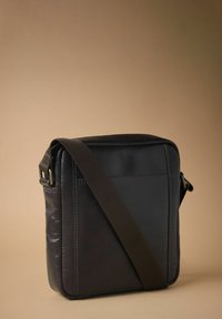 Dark leather messenger bag with a square shape, adjustable brown strap, and minimalistic design; features a back pocket and smooth texture.