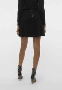 Jupe skater noire avec un ourlet évasé, détail de fermeture éclair à l'arrière, associée à des bottines à talons noires, texture lisse.